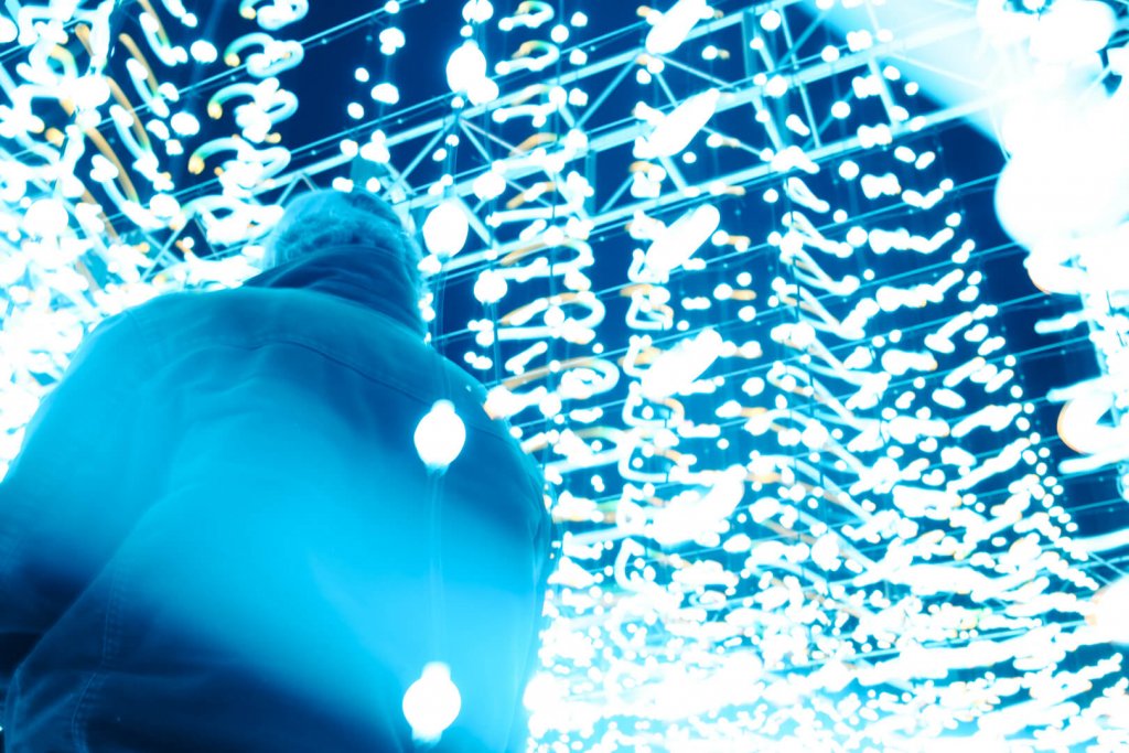 A photograph of a single man from a low angle, standing under a set of lights on strings with a blue glow